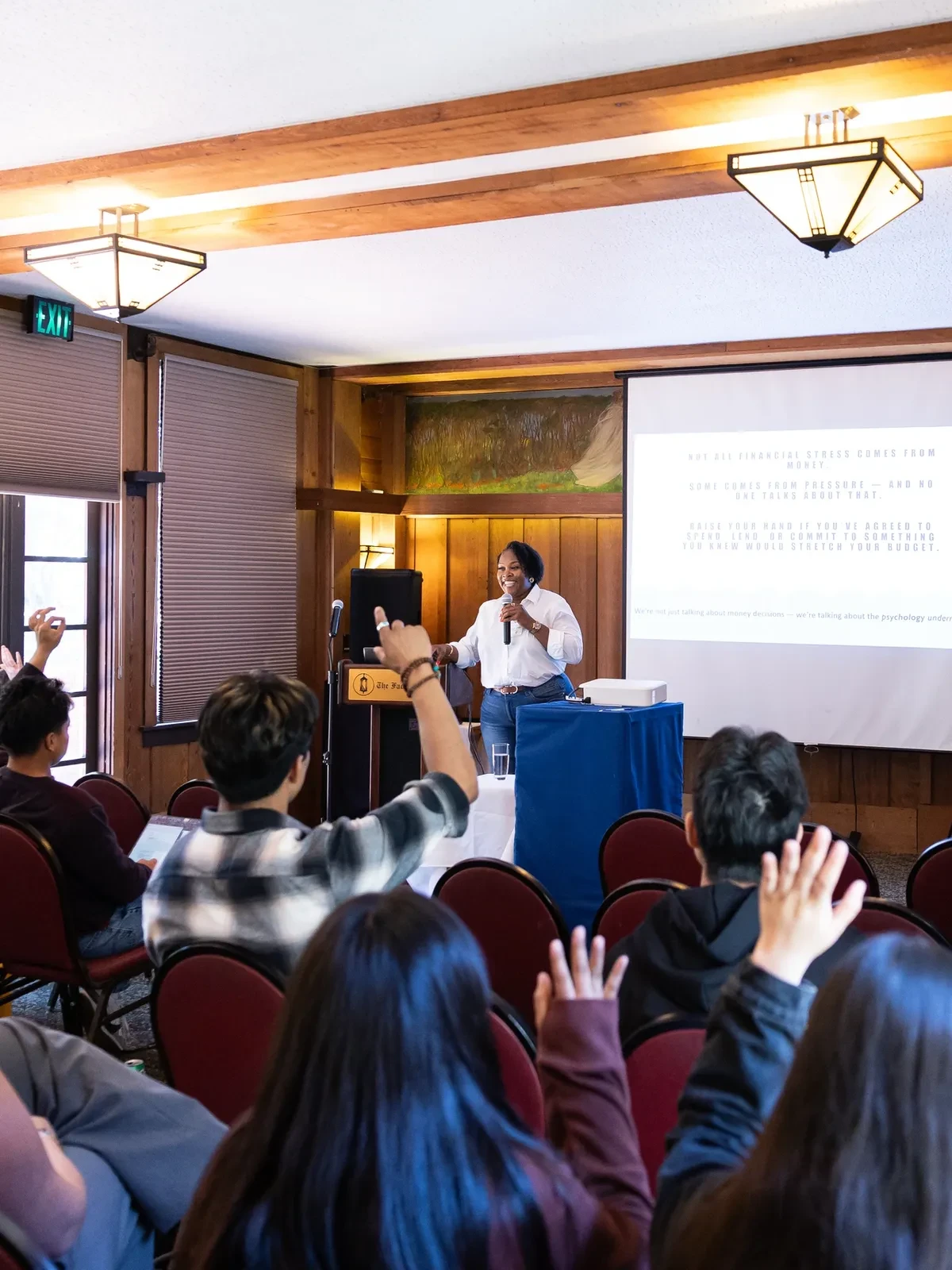 Ockemia Bean speaking at a podium in a wood-paneled venue with audience members raising hands